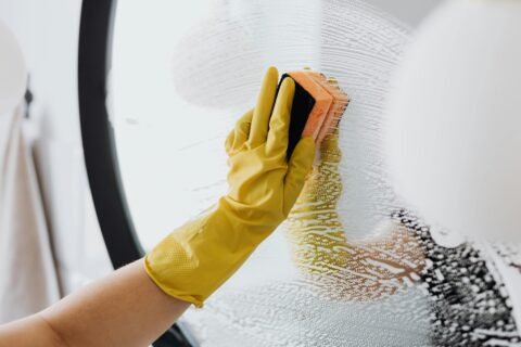 A person wearing yellow gloves meticulously cleans a window with a scrub brush, ensuring a streak-free shine.